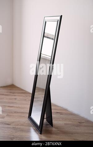 a rectangular mirror in a brown wooden frame stands indoors on a white background, the mirror is in an empty room Stock Photo