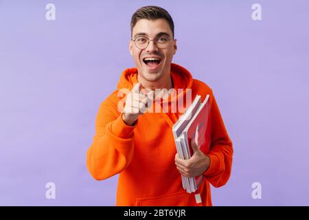 Photo of laughing positive young man student posing isolated over purple background holding copybooks pointing to you. Stock Photo