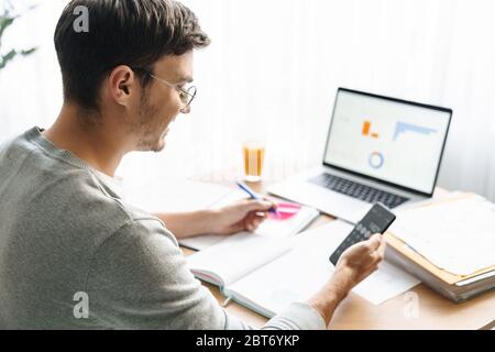 Image of happy young man in eyeglasses doing calculations while working on laptop at home Stock Photo