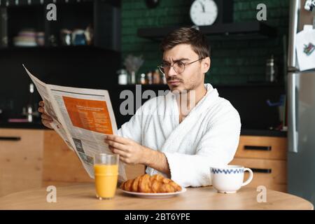 Upset young man wearing bathrobe having breakfast while sitting in the kitchen and reading newspaper in the morning Stock Photo