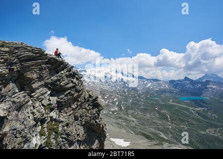 Beautiful view of traveler sitting on the edge of high rocky cliff under cloudy sky. Man tourist admiring view of mountain valley with hills and blue lake. Concept of travelling, hiking and alpinism. Stock Photo