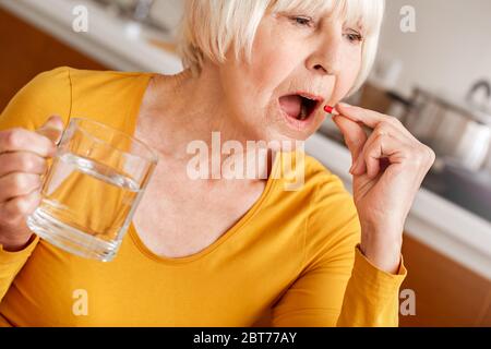 Retirement. Senior woman sitting at table at kitchen with cup of water and pill coronavirus treatment close-up blurred background Stock Photo