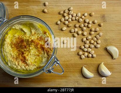 Homemade hummus in glass jar with chickpea beans and garlic cloves on wood background Stock Photo