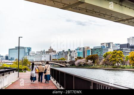 Osaka, Japan - April 13, 2019: Okawa river water view of cityscape downtown skyline under bridge in spring and people walking in park Stock Photo