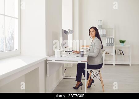 Beautiful busy serious business woman in a gray jacket looks sitting at a workplace at a table in a white office. Stock Photo