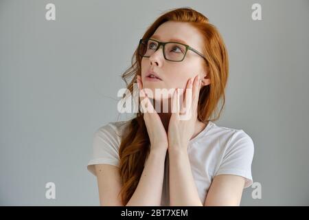 A red-haired girl with glasses stands on a gray background and looks to the top with her palms to her face Stock Photo