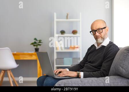 Work online at home. Serious middle aged man gray beard with laptop working online sitting in an armchair in a room. Stock Photo