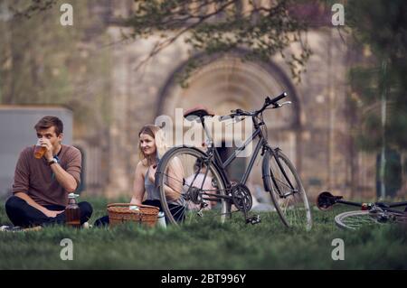 A white young couple having picnic in a park with vintage bicycles, picnic basket and drinking beer from plastic cap Stock Photo