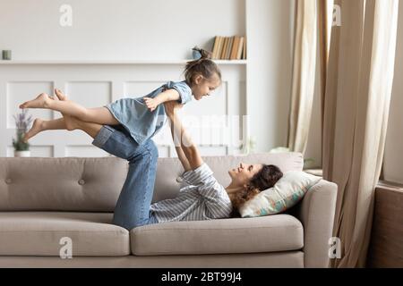 Smiling mother holding little daughter pretending flying, lying on couch