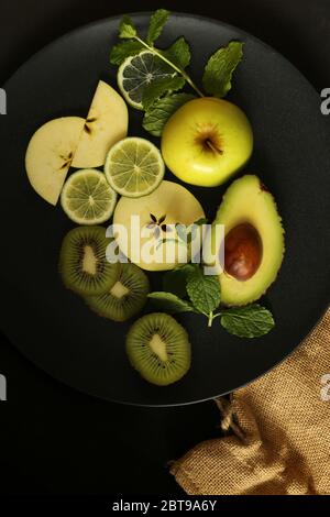Multiple green fruits on round black plate on black background with canvas. Dramatic dark food.Flat lay, close up, top view Stock Photo