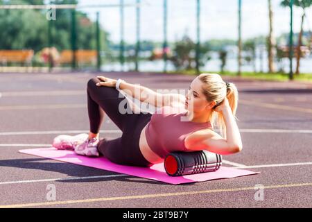 Motivated sporty woman training on mat outdoor summer day, using foam roller massager for relaxation, stretching spine muscles, doing fascia exercise Stock Photo