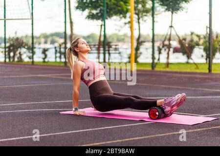 Athletic blond sporty woman training on mat outdoor summer day, massaging and stretching hamstring and ankle leg muscles on foam roller, performing fa Stock Photo