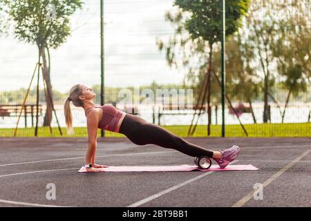 Fit young woman training on mat outdoor summer day, performing reverse plank with foam roller massager, stretching spinal muscles, doing fascia exerci Stock Photo