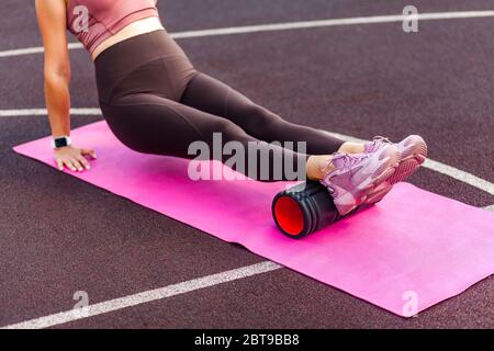 Closeup sporty woman training on mat outdoor summer day, massaging and stretching hamstring and ankle leg muscles on foam roller, performing fascia ex Stock Photo