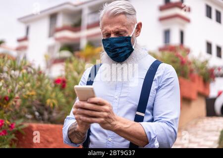 Senior hipster man using smart mobile phone outdoor while wearing face safety mask - Mature guy watching video on smartphone during coronavirus outbre Stock Photo