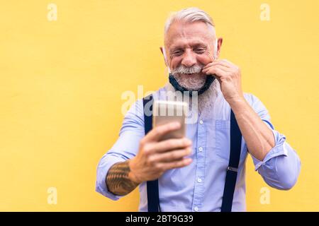 Trendy hipster man using mobile phone doing video call while wearing fashion silk safety mask - Outfit, technology and lifestyle during coronavirus ti Stock Photo