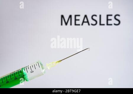 This photo illustration shows a disposable syringe with hypodermic needle, MEASLES written on a white board behind Stock Photo