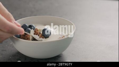 man hand tastes chocolate granola with almond flakes and blueberries in white bowl on concrete surface Stock Photo
