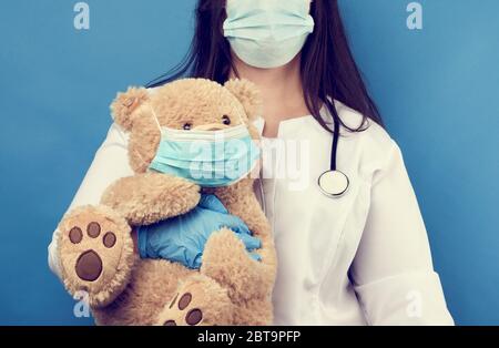 woman doctor pediatrician holds brown teddy bear in hand in white medical disposable mask, concept of preventing epidemics and pandemics against flu Stock Photo
