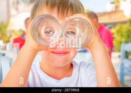 Funny kid looking through two drink glasses as if it were binoculars. Child having fun while waiting for order at cafe, restaurant. Happiness and Stock Photo