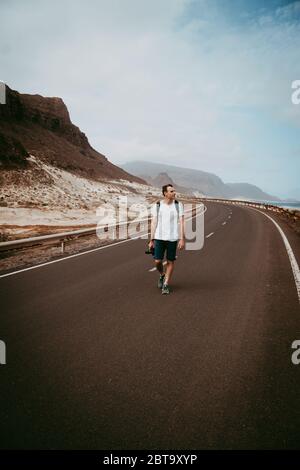 Traveler walks in the center of an epic winding road. Huge volcanic mountains in the distance behind him. Sao Vicente Cape Verde Stock Photo