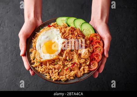 Nasi Goreng - Indonesian Chicken Fried Rice on black plate in female hands. Nasi Goreng is an Indonesian cuisine dish with jasmine rice, chicken meat, Stock Photo