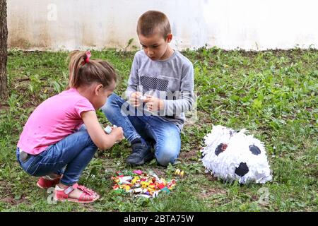 Two children eat sweets candy on the ground in the garden from a broken pinata toy. Girl and boy squat on the grass and unpacking candies Stock Photo