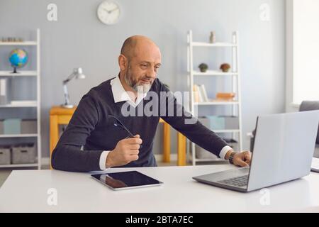 An elderly man works using a laptop office at home. Mature confident man with a gray beard analyzes a business strategy remotely leads a startup sitti Stock Photo