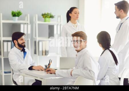 Male smiling doctor sitting at a table with collegues in white lab coats at a medical hospital. Confident doctors at the conference. Stock Photo