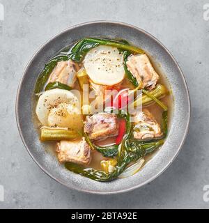 Sinigang na Baboy or Filipino Pork Meat Soup in gray bowl on concrete backdrop. Sinigang is a Filipino cuisine dish with meat, bamia, daikon, spinach, Stock Photo