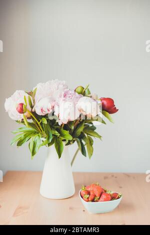 Toned photo Bunch of Pink peonies in vase and strawberry on the wooden table Flowers on a beige wooden table near the window. Home house interior Stock Photo