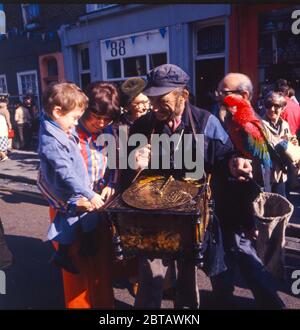 A street busker with his music box and parrot entertain a young boy & the crowds in Portobello Road antique market in Notting Hill, London in the 1970s. Stock Photo