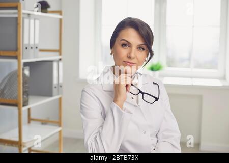 Smart adult woman in white robe taking off glasses and looking at camera while working in light office of contemporary clinic Stock Photo