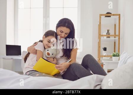 Happy Mother's Day. Mother and daughter are reading a book while sitting on the bed at home. Mom reads an interesting story to her child in a book bef Stock Photo