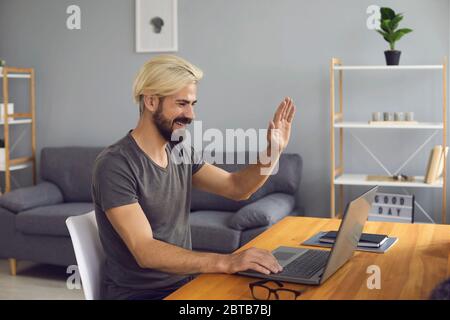 Happy bearded man smiling and waving hand while sitting at table and making video call to colleague at home Stock Photo