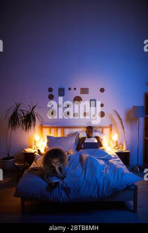 Vertical wide angle portrait of young couple using computer gadgets in bed at night, copy space Stock Photo