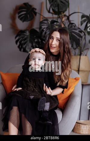 Beautiful mother sits on a chair and keeps her daughter on lap at home. family look concept. mother's, baby's day Stock Photo
