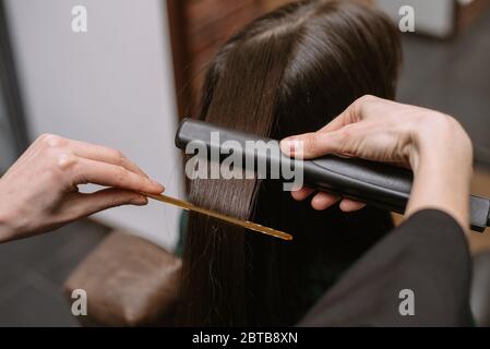 The hairdresser pulls and straightens the hair of a brunette in a beauty salon with a comb and ironing. Keratin recovery hair and protein treatment pi Stock Photo