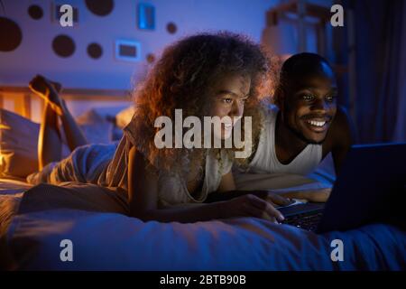 Portrait of young mixed-race couple using laptop and smiling while lying in bed at night, copy space Stock Photo