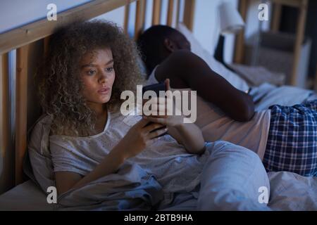High angle portrait of young curly-haired woman using smartphone in bed at night while suffering from insomnia, copy space Stock Photo