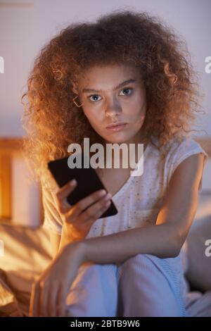 Vertical portrait of young curly-haired woman using smartphone while sitting on bed at night and looking at camera Stock Photo