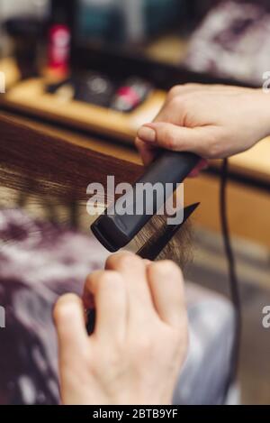 The hairdresser pulls and straightens the hair of a brunette in a beauty salon with a comb and ironing. Keratin recovery hair and protein treatment pi Stock Photo