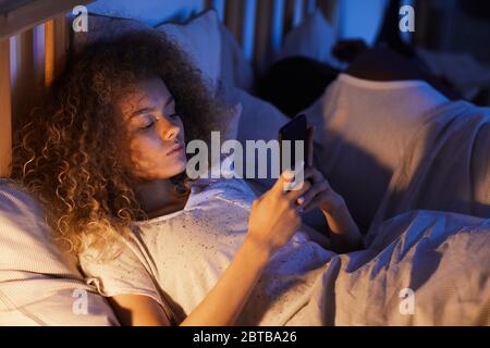 High angle portrait of curly-haired young woman using smartphone in bed at night while lying next to husband or boyfriend, copy space Stock Photo