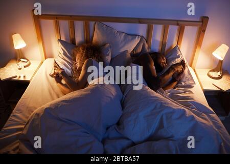 Above view portrait of young mixed-race couple lying back to back in bed and using smartphones, copy space Stock Photo
