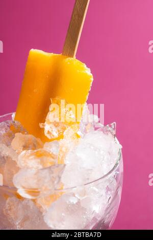 Ice cream popsicle close-up with orange flavor, on a pink background. Ice cream on stick in a bowl with crushed ice. Refreshing summer fruity dessert. Stock Photo