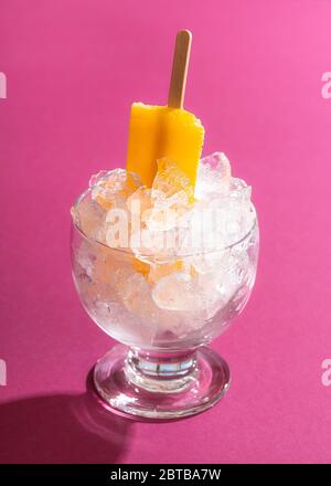 Orange ice cream popsicle in a bowl with crushed ice on a pink seamless background in harsh light. Cooling summer dessert. Vegan ice cream on stick. Stock Photo