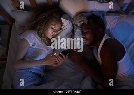 Above view at young mixed-race couple lying in bed at night and using smartphones, copy space Stock Photo