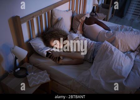 High angle portrait of curly-haired young woman using smartphone while lying with boyfriend in bed at night, copy space