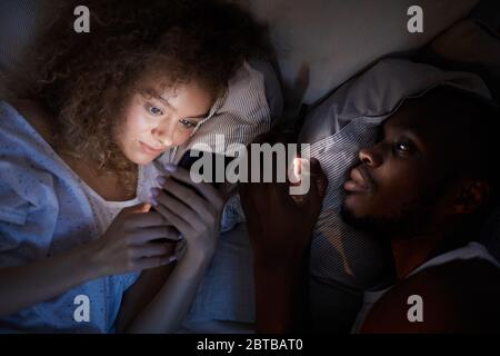 Above view at young mixed-race couple using smartphones while lying in bed at night, focus on smiling curly-haired woman Stock Photo