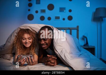 Portrait of carefree mixed-race couple looking at smartphone screen and smiling while lying in bed at night, copy space Stock Photo